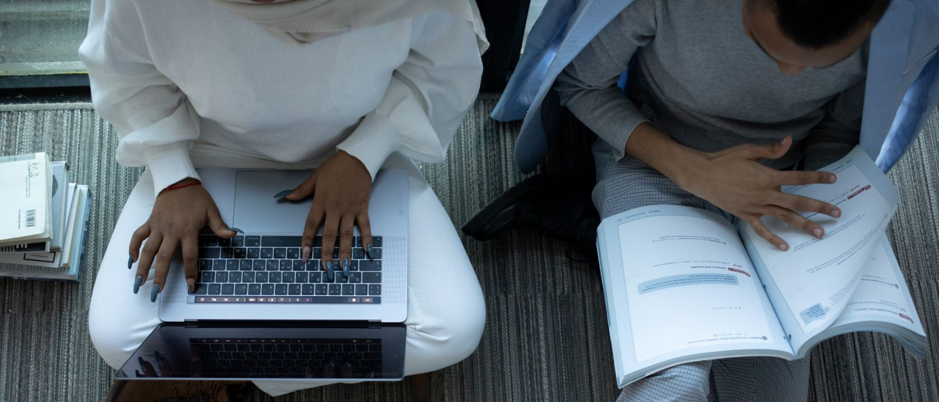 Zwei Menschen sitzen auf dem Boden, eine schaut in ihren Laptop, der andere ließt ein Heft