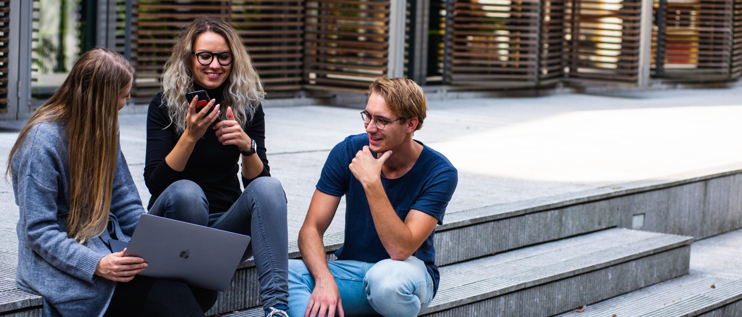 Drei junge Menschen sitzen mit Laptop und Handy auf einer Treppe und unterhalten sich