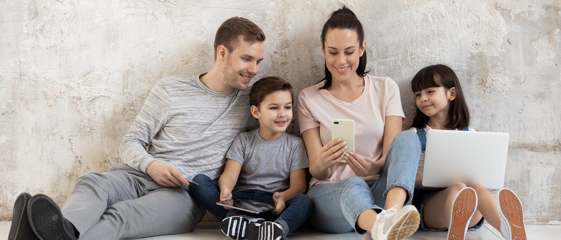 Vater, Mutter und zwei Kinder sitzen auf dem Boden und schauen gemeinsam in ein Handy