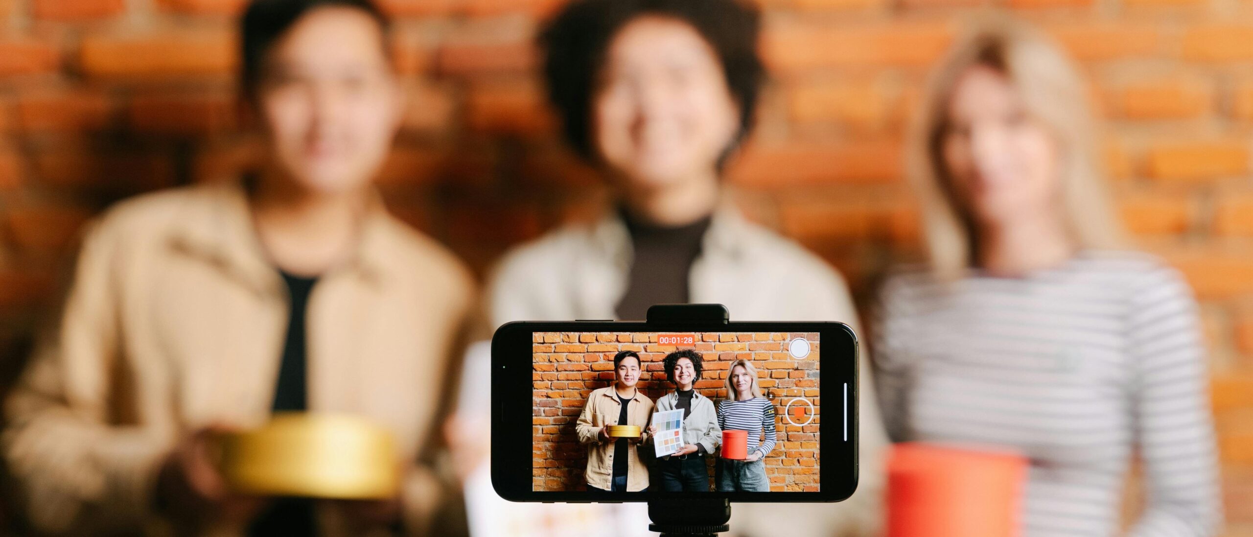 Eine Handykamera, die gerade drei Menschen abfotografiert. Im Hintergrund sind die Menschen zu sehen.