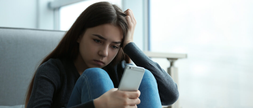 Mädchen sitzt mit angewinkelten Beinen vor ihrem Handy und schaut verzweifelt in das Display
