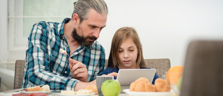 Vater sitzt mit seiner Tochter vor dem Tablet und erklärt ihr die Funktionen.