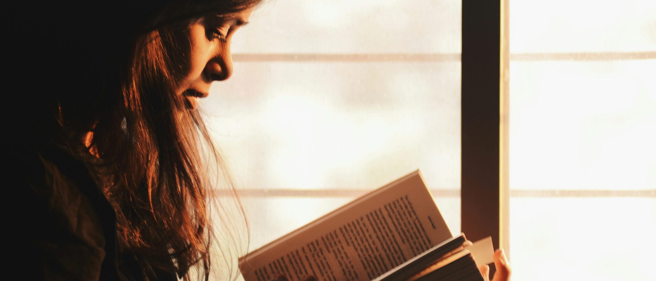 Mädchen sitzt vor einem Fenster und liest gebannt in ihrem Buch.