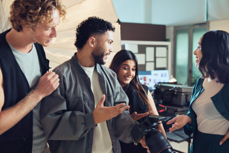 Vier Menschen tauschen sich stehend aus. Eine Frau in der Mitte hält eine Kamera und schaut das Bild darauf an.