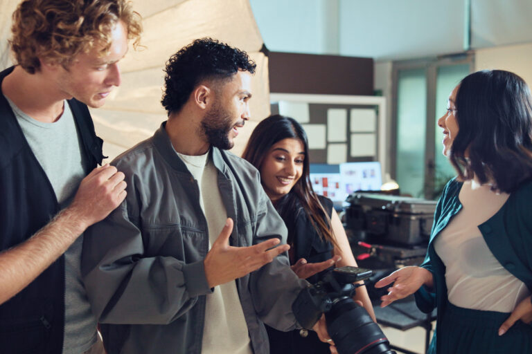Vier Menschen die sich austauschen. Eine von Ihnen hält eine Kamera in der Hand und schaut sich das Bild darauf an.