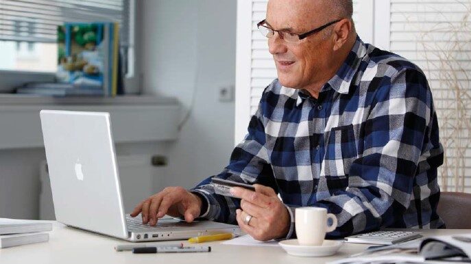 Ein älterer Herr sitzt vor seinem Laptop und hält eine Kreditkarte in der Hand.