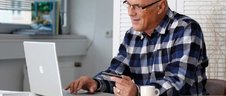 Ein älterer Herr sitzt vor seinem Laptop und hält eine Kreditkarte in der Hand.
