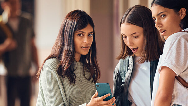 In einem Schulgang sind drei Jugendliche um ein Handy versammelt und schauen entsetz in Richtung einer Person